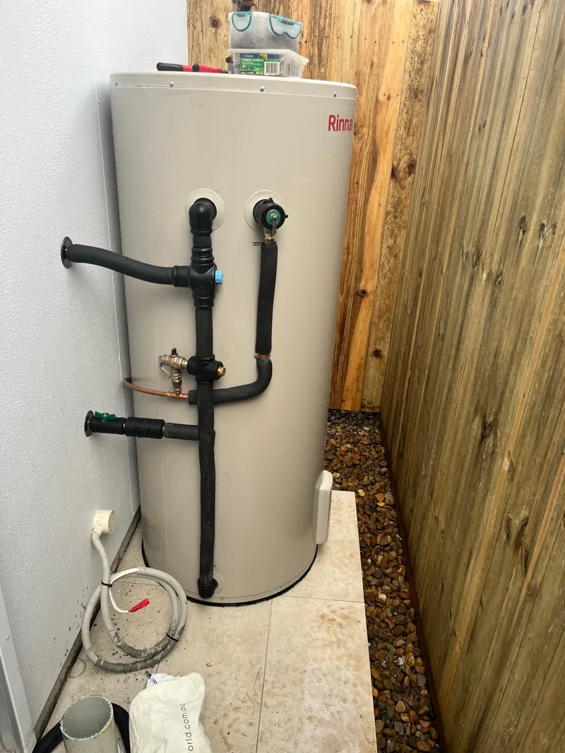 A newly installed Rinnai electric hot water storage tank with black pipework against a timber fence