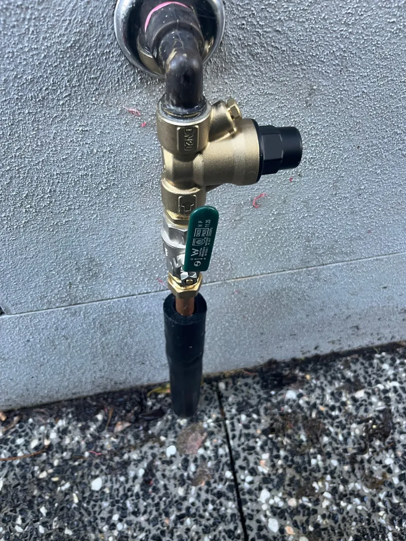 A brass tempering valve fitted to hot water pipework with a black relief drain line running to the ground